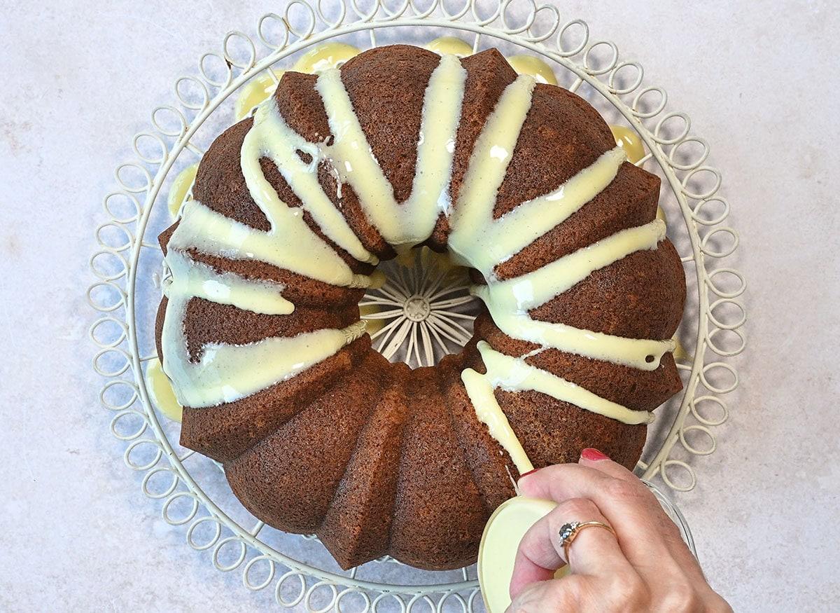 A banana bundt cake on a decorative wire rack is being drizzled with white icing by a hand using a small jug. The cake has an elegant swirl pattern and the icing is spread across the top in wavy lines.