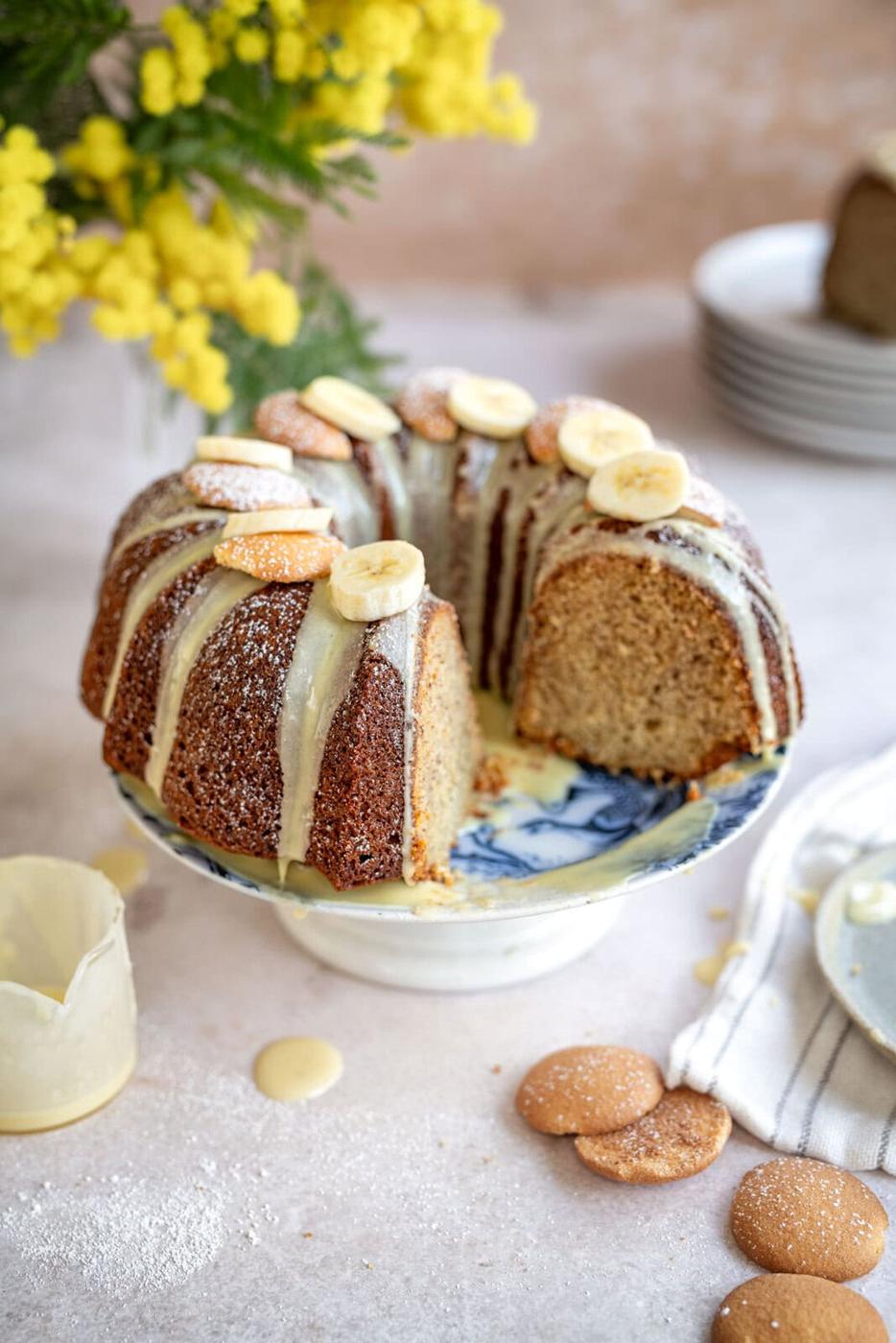 A bundt cake with vanilla glaze is displayed on a blue and white cake stand. The cake is topped with banana slices and cookies. A slice is cut out, revealing the moist interior. Yellow flowers and a stack of plates are in the background.