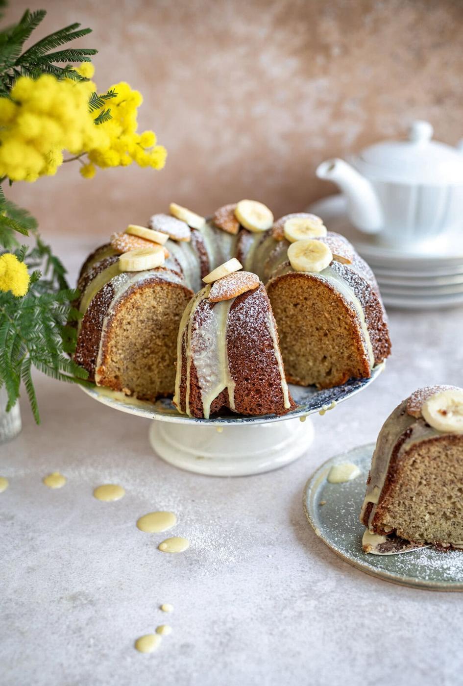 Banana pudding cake with sliced bananas and icing drizzled on top, displayed on a white cake stand. A slice is served on a small plate beside it. Yellow flowers and a white teapot are in the background on a light-colored surface.
