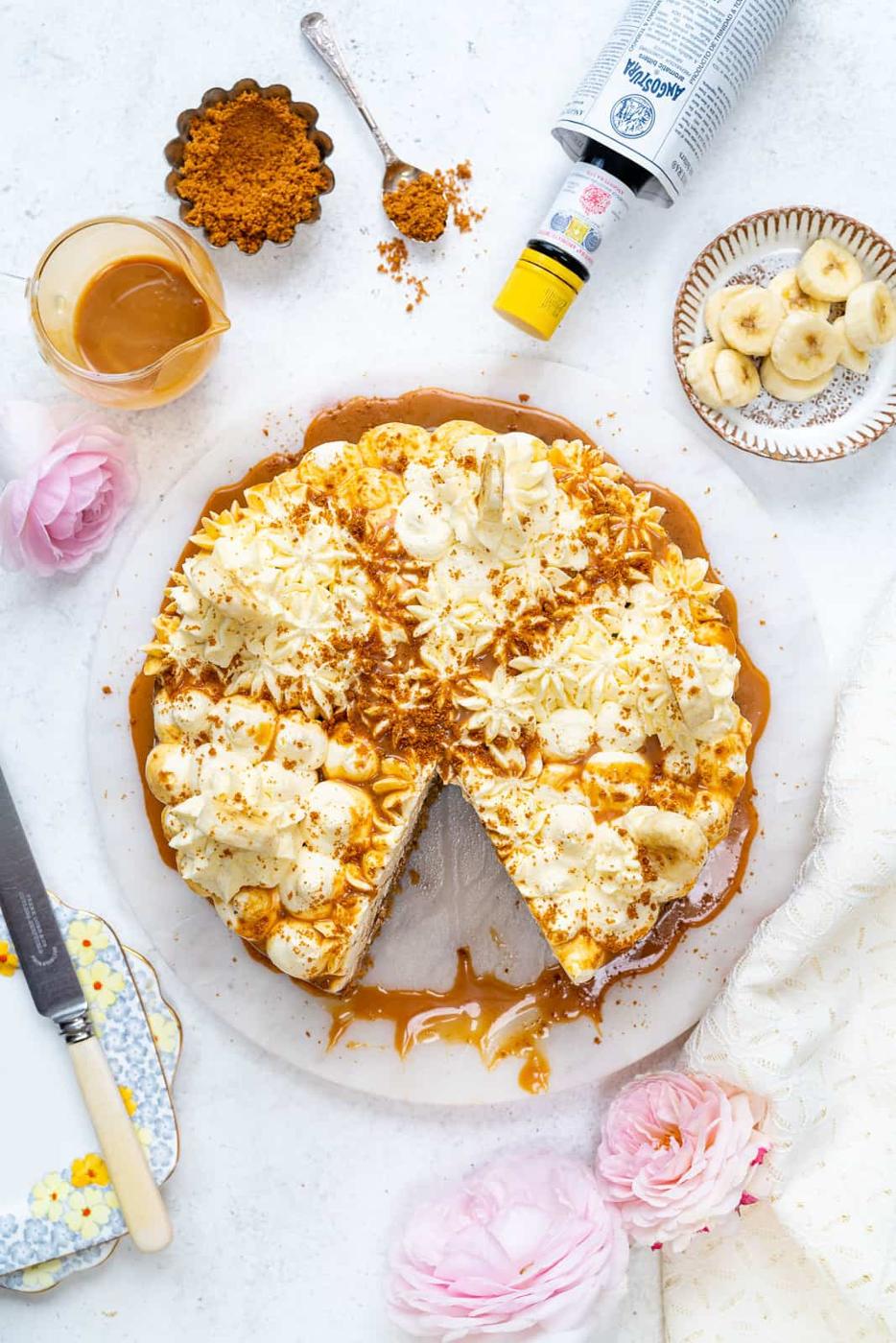 A Banoffee Cake sits on a white surface, topped with whipped cream, banana slices, and biscuit crumbs. A slice is missing. Nearby are caramel sauce, more banana slices, biscuit crumbs, whipped cream, plates, and pink flowers.