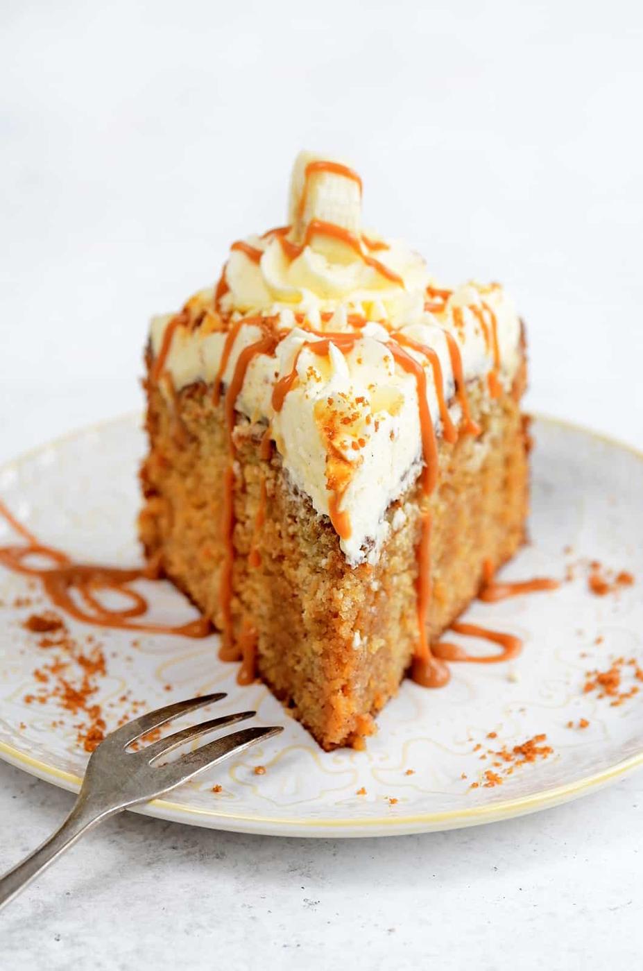 A slice of moist Banoffee Cake topped with swirls of whipped cream, banana slices, and a drizzle of caramel sauce sits on a white plate, with a fork beside it.