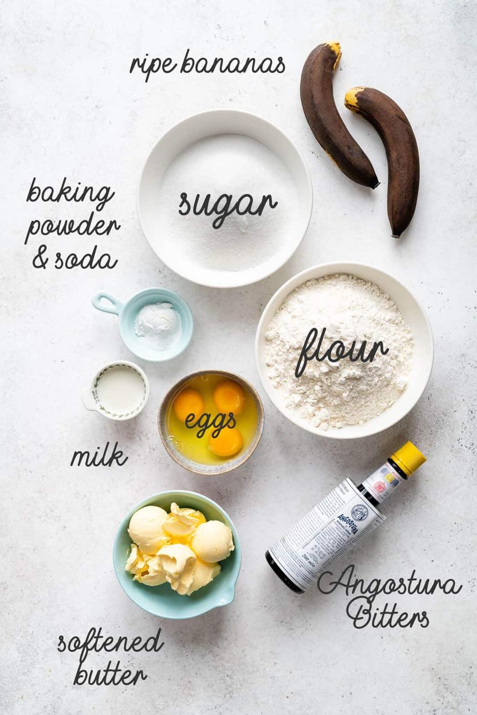 A flat lay of ingredients for banana bread or Banoffee Cake: two ripe bananas, a bowl of sugar, a bowl of flour, two eggs, softened butter, baking powder & soda, milk, and a bottle of Angostura bitters—all labeled.