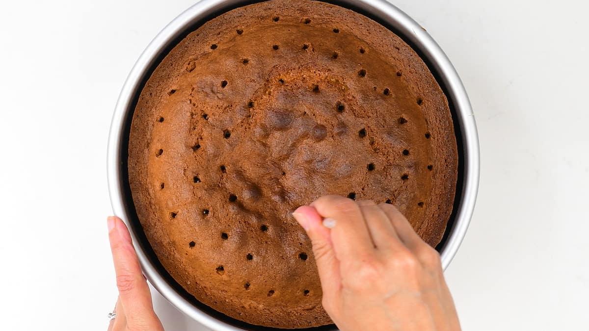 A person uses a skewer to poke holes in the top of a round, golden-brown Banoffee Cake still in its baking pan, photographed from above.