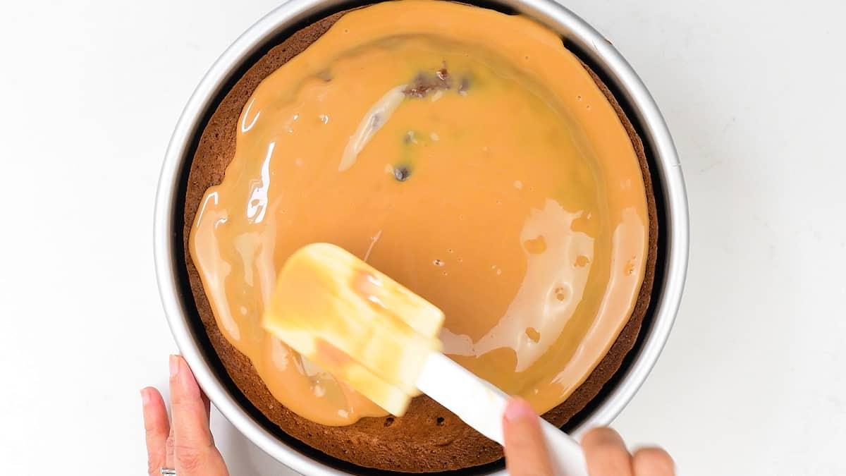 A hand uses a spatula to spread caramel-colored glaze over a round Banoffee Cake in a metal pan on a white surface.