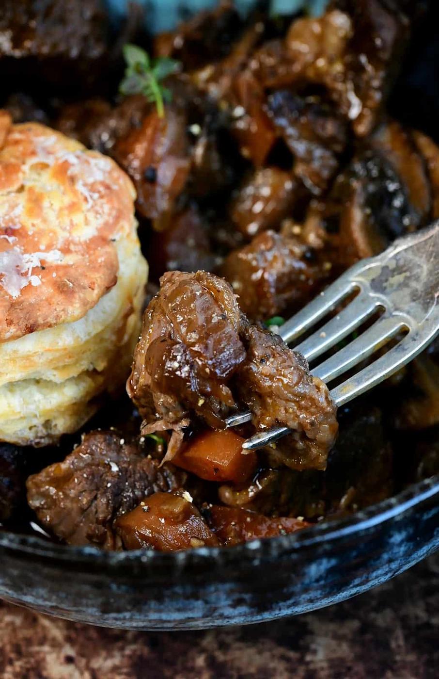 Close-up of a fork holding a piece of tender beef stew with carrots, next to a flaky biscuit, all served in a dark bowl—an inviting take on classic beef cobbler.