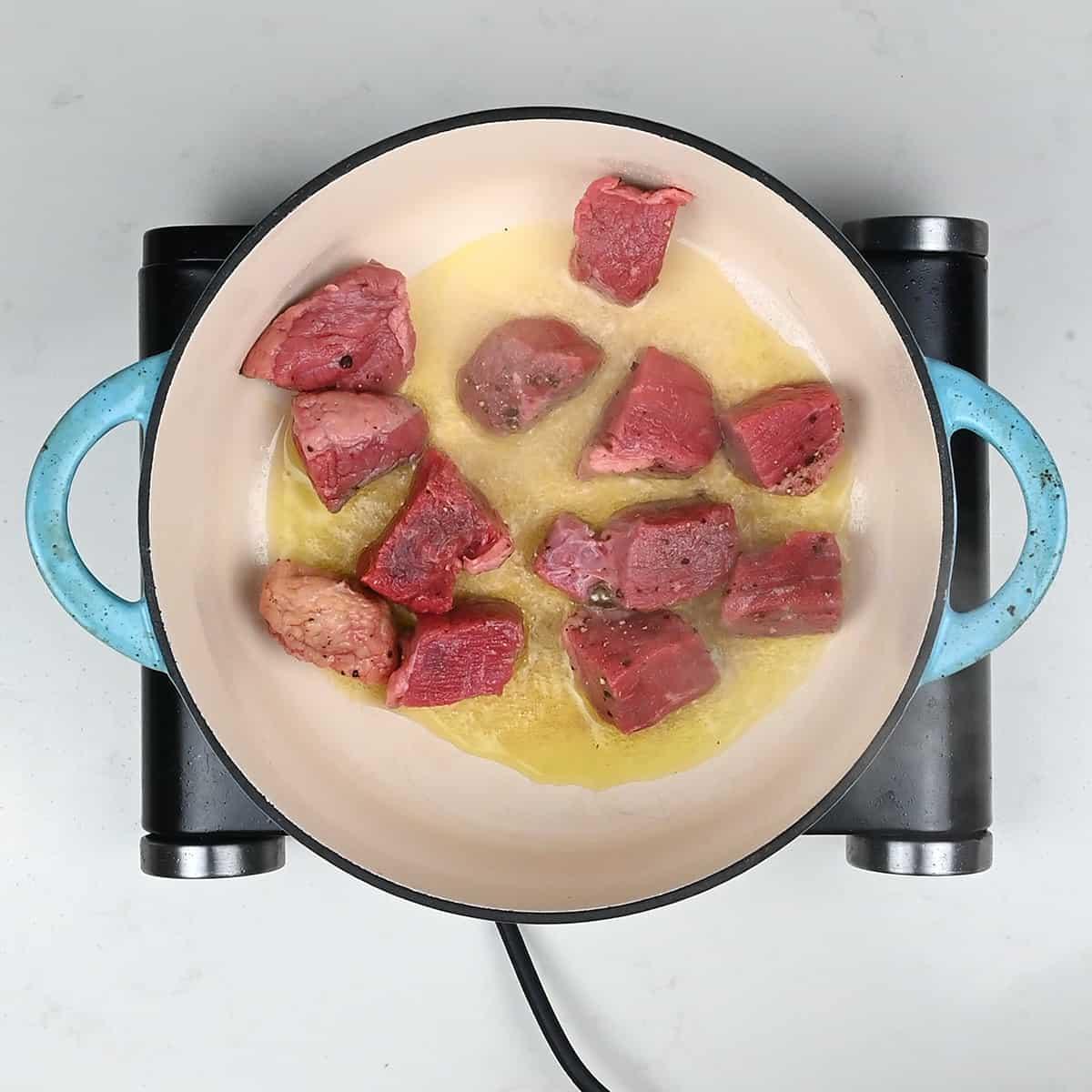 Chunks of raw beef for a hearty beef cobbler are being seared in melted butter inside a round, white pot with blue handles, set on an electric stovetop.