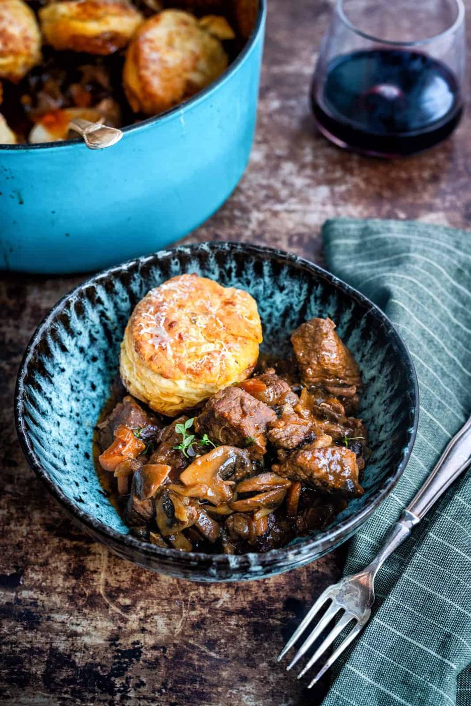 A bowl of beef cobbler with vegetables, topped with a golden biscuit, sits next to a green napkin with a fork. In the background, a blue pot and a glass of red wine rest on a rustic wooden surface.