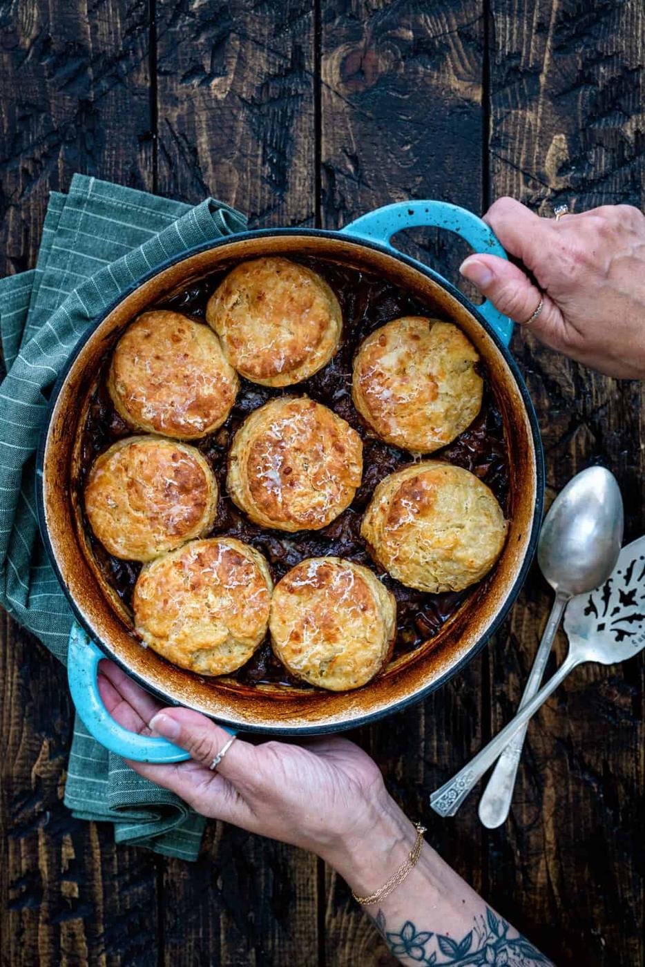 A person holds a round blue baking dish filled with golden-brown biscuits atop a savory beef cobbler. The dish rests on a green cloth beside two vintage serving utensils on a rustic wooden table.
