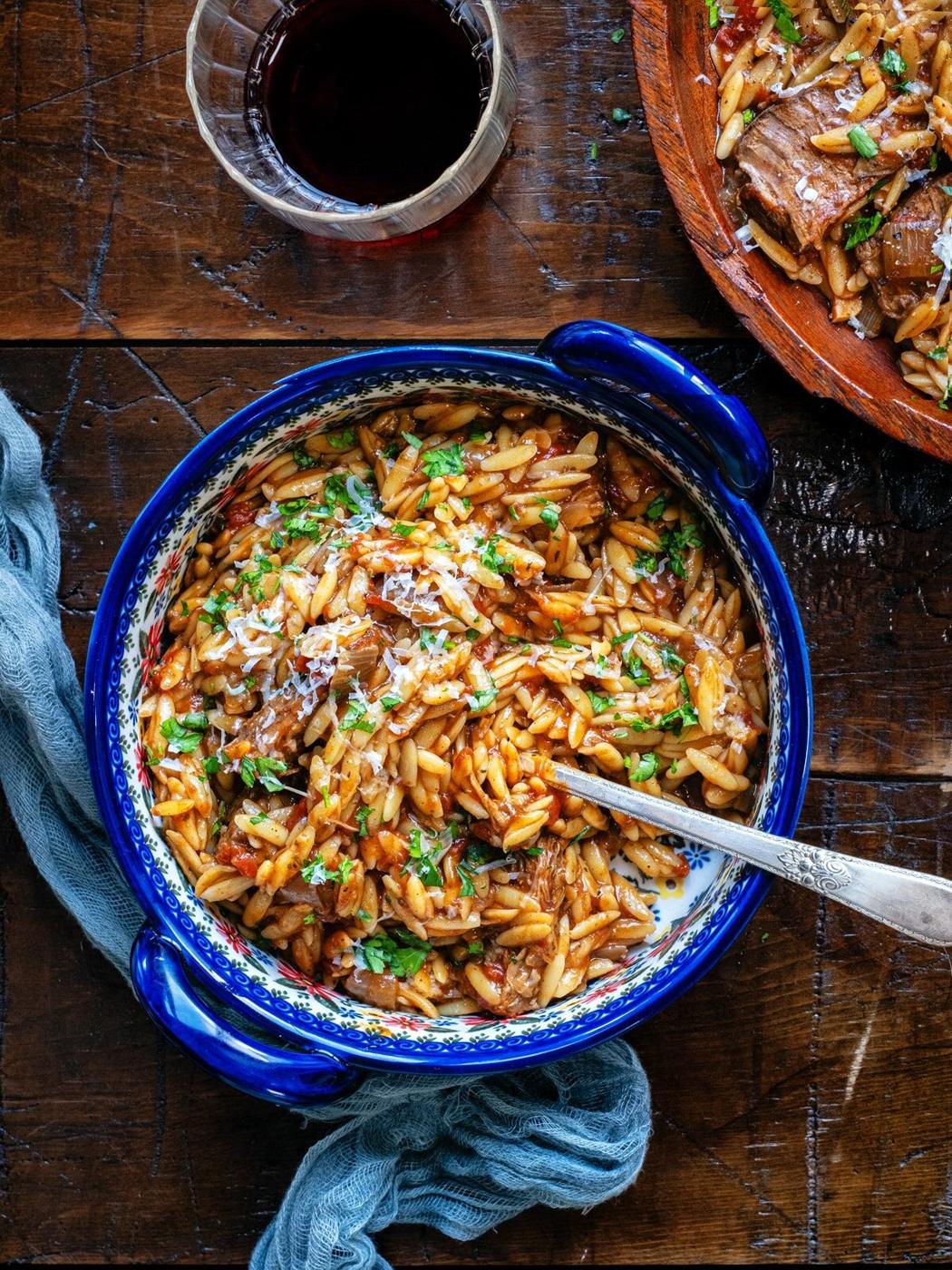 A blue bowl filled with orzo pasta in a tomato-based sauce, reminiscent of Greek Beef Giouvetsi, is topped with grated cheese and fresh herbs. It sits on a wooden table beside a glass of red wine and a blue cloth napkin.
