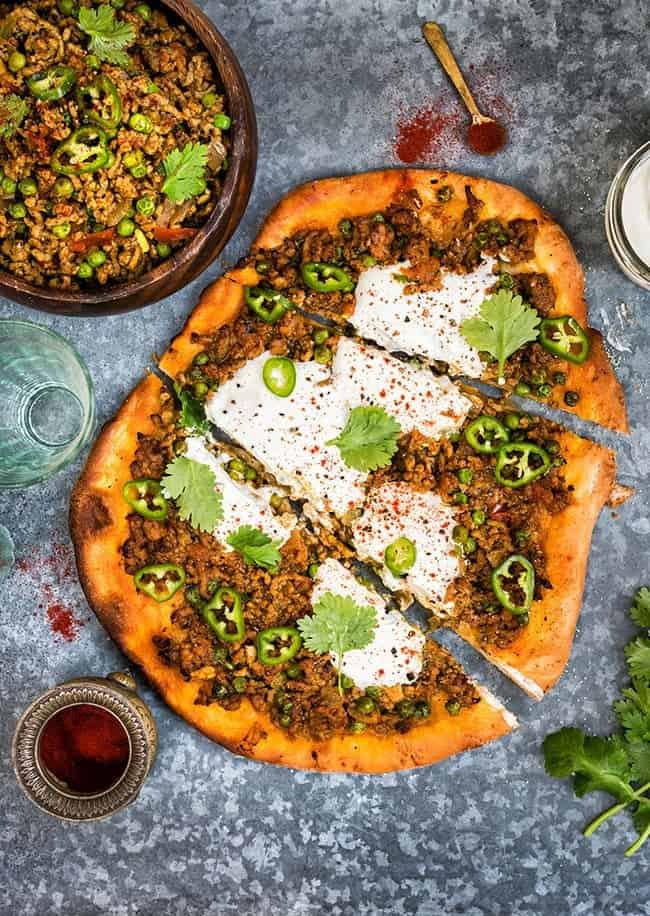 A flatbread topped with ground beef Keema Curry, green peas, sliced green chilies, yogurt sauce, and cilantro. Its served on a dark textured surface beside a bowl of the savory meat mixture and surrounded by fresh herbs and spices.