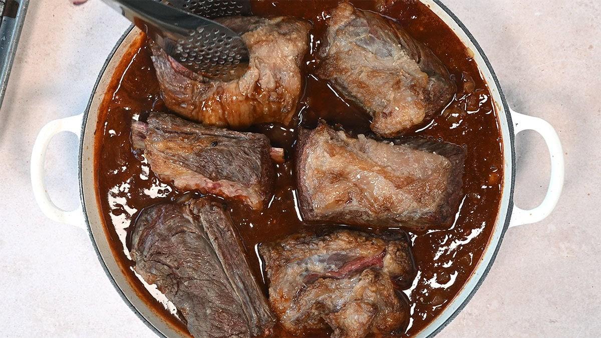 adding seared short ribs into a skillet with French onion red wine braise