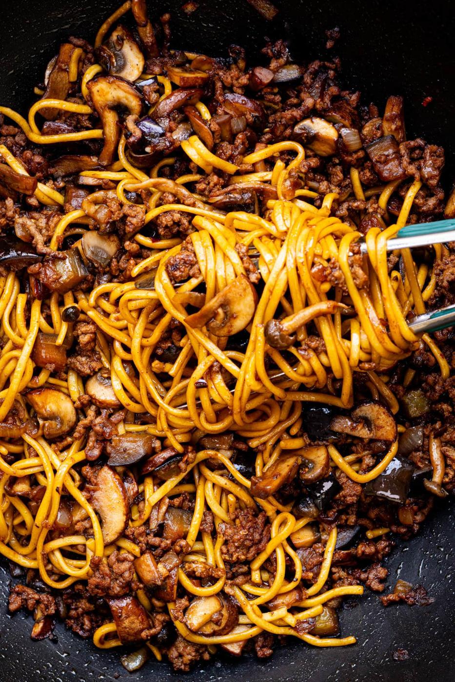 Chopsticks lifting a portion of stir-fried noodles mixed with ground beef, sliced mushrooms, onions, and a dark savory sauce in a pan.
