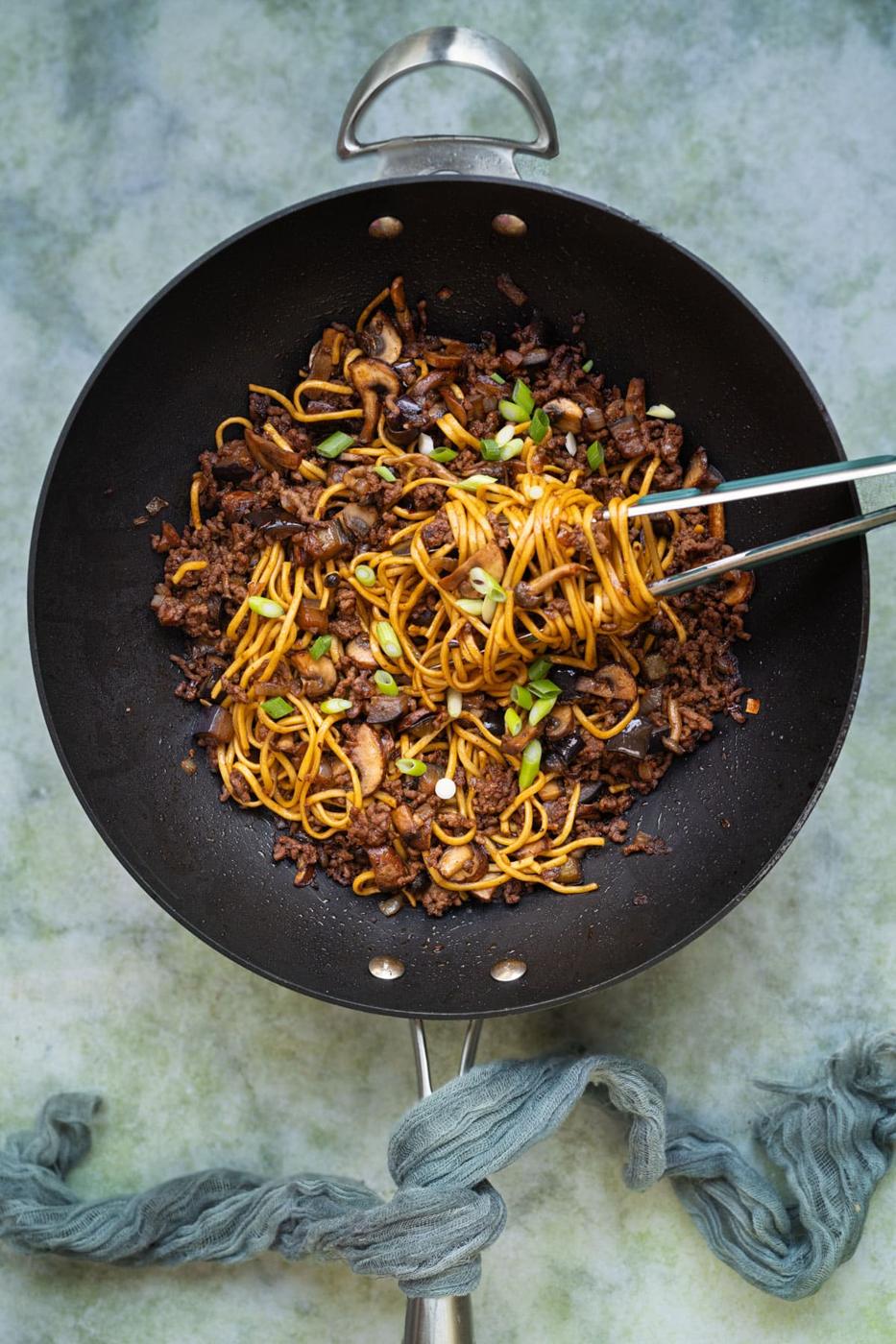A black wok filled with stir-fried noodles, mushrooms, ground meat, and chopped green onions. A pair of metal tongs is lifting a portion of noodles. The wok sits on a textured green surface with a blue-grey cloth nearby.