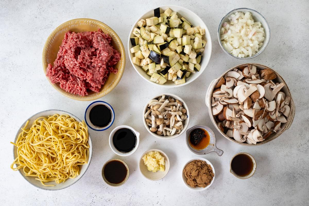 Bowls containing raw ground beef, chopped eggplant, diced onions, sliced mushrooms, mixed mushrooms, yellow noodles, minced garlic, brown sugar, soy sauce, oyster sauce, and other sauces on a white surface.