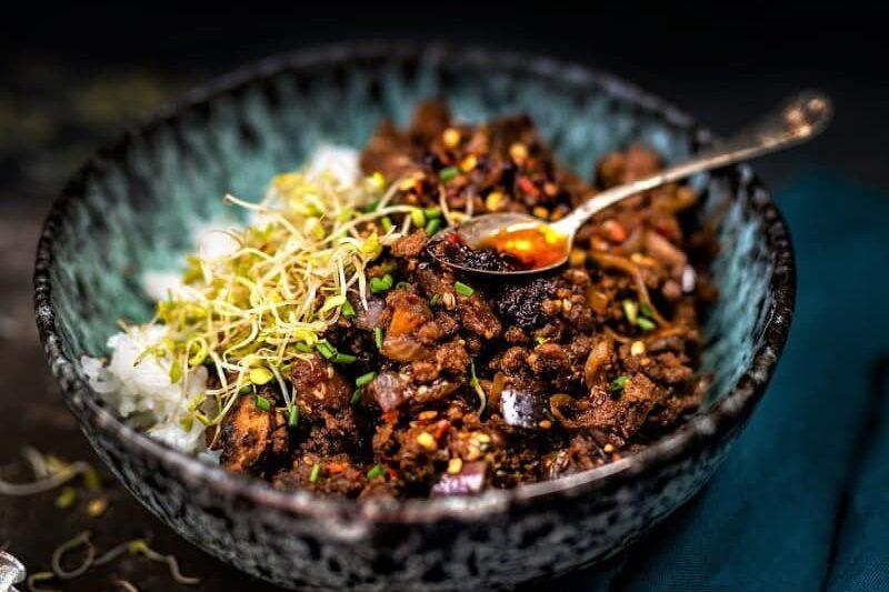 A ceramic bowl filled with rice, topped with spiced ground beef stir fry, fresh sprouts, and chopped herbs. A small spoon rests in the bowl, which sits on a dark surface with a teal cloth nearby.