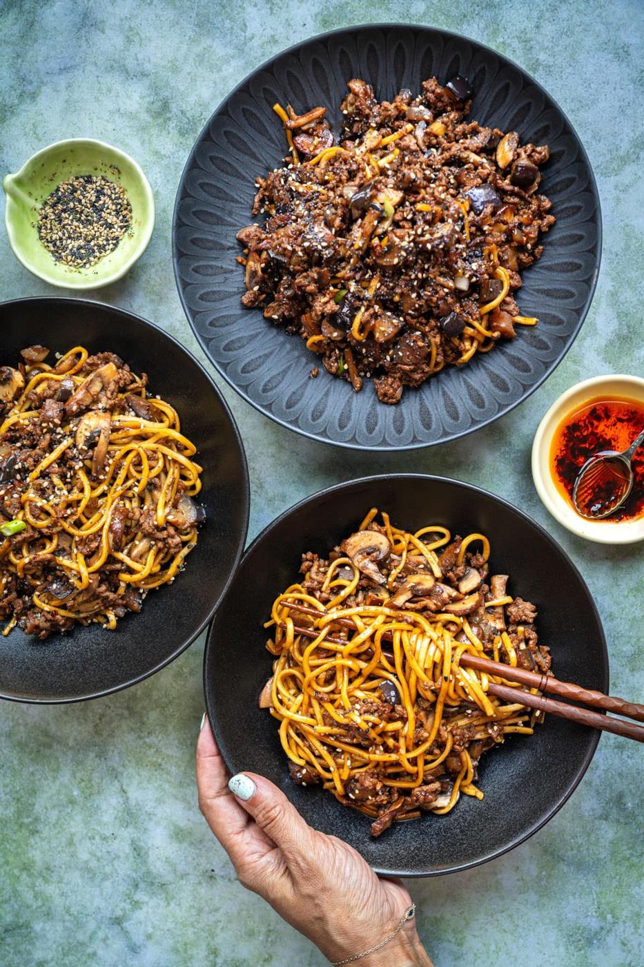 Three bowls of noodles with ground beef, mushrooms, and sesame seeds are arranged on a table, with chopsticks in one hand. Small dishes of chili oil and sesame seeds are placed beside the bowls.