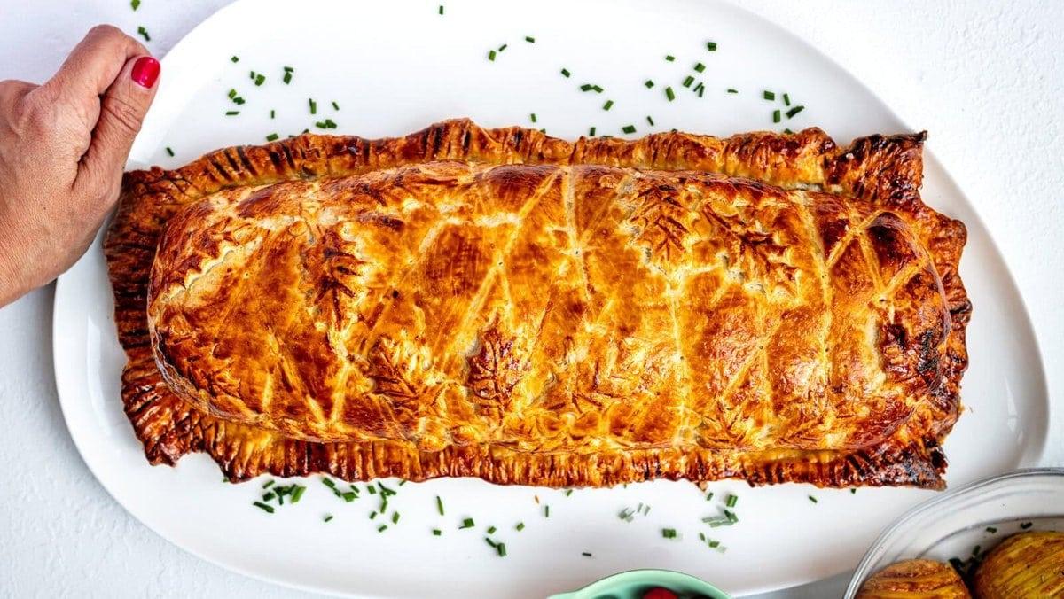 A golden-brown baked pastry with decorative patterns rests on a white oval platter, garnished with chopped chives. Nearby are a bowl of cranberries and sliced roasted potatoes. A hand holds the platter edge.