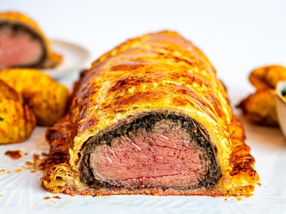 A sliced beef Wellington shows layers of flaky golden pastry, mushroom duxelles, and medium-rare cooked beef tenderloin on a white plate, with more slices and side dishes in the background.