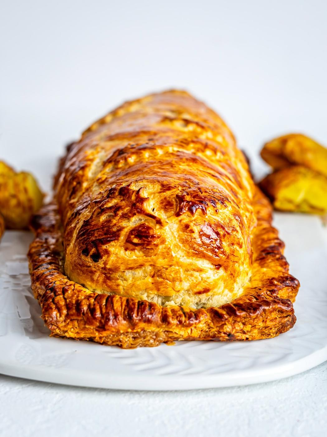 A golden-brown, baked pastry loaf with a flaky crust sits on a white plate, with a few roasted potatoes in the background.