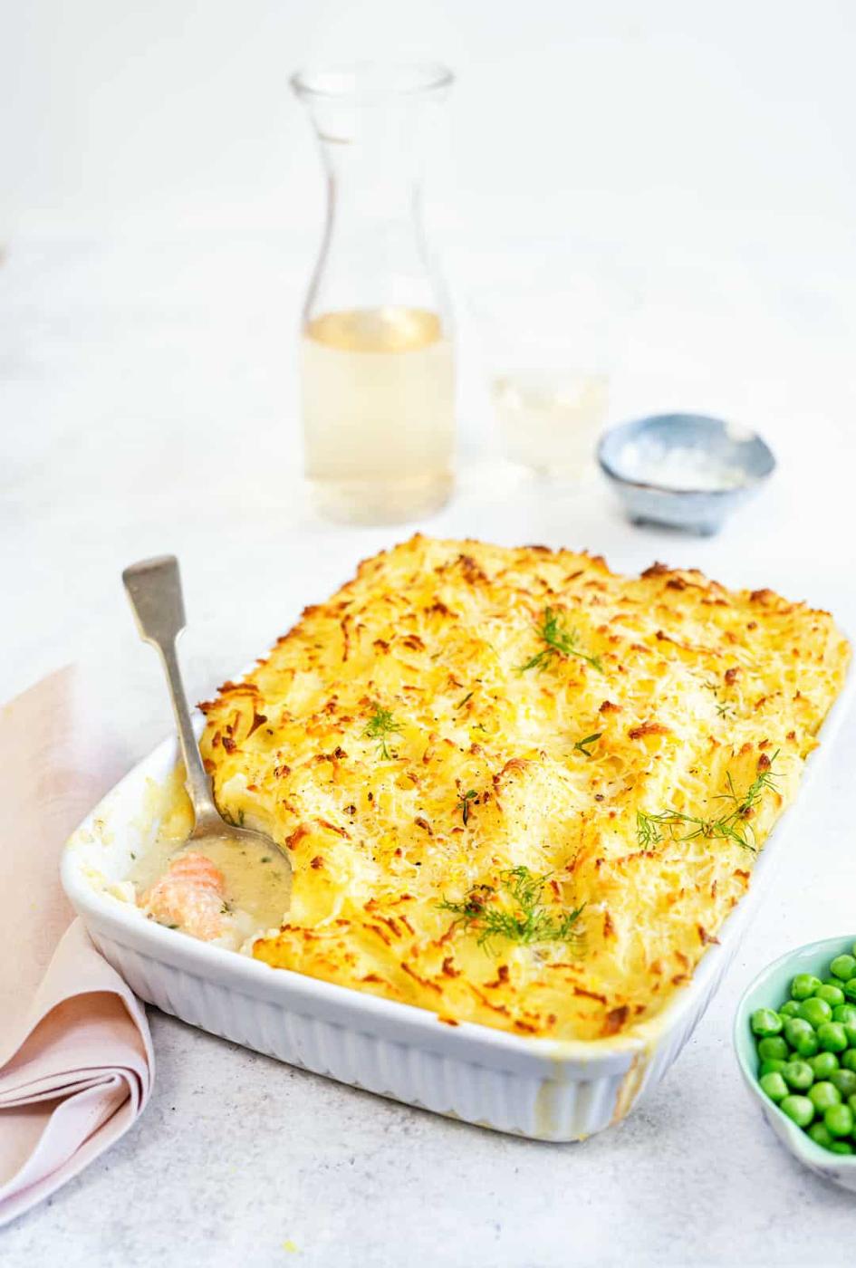 Fish pie with a crisp cheesy mashed potato topping in a white dish, with a serving spoon and a bowl of peas