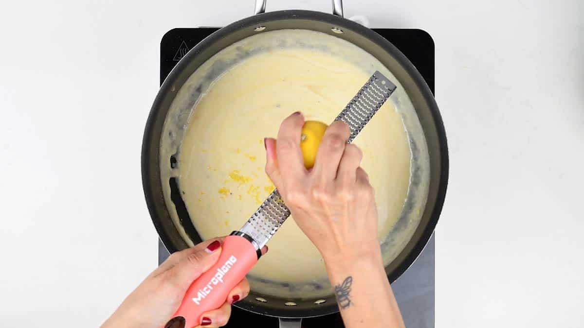 Lemon zest being grated over the creamy fish pie sauce in a pan