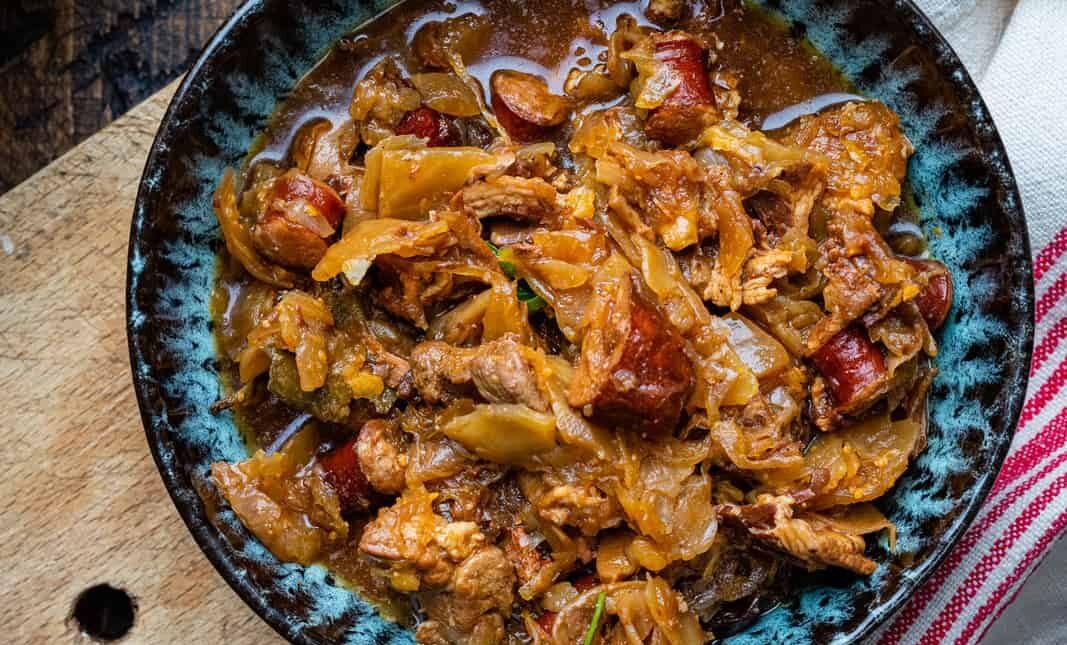 A bowl of hearty Bigos, or Polish Hunter's stew, featuring shredded cabbage, chunks of sausage, and pieces of pork in a rich brown sauce, served on a wooden surface with a striped cloth partially visible.
