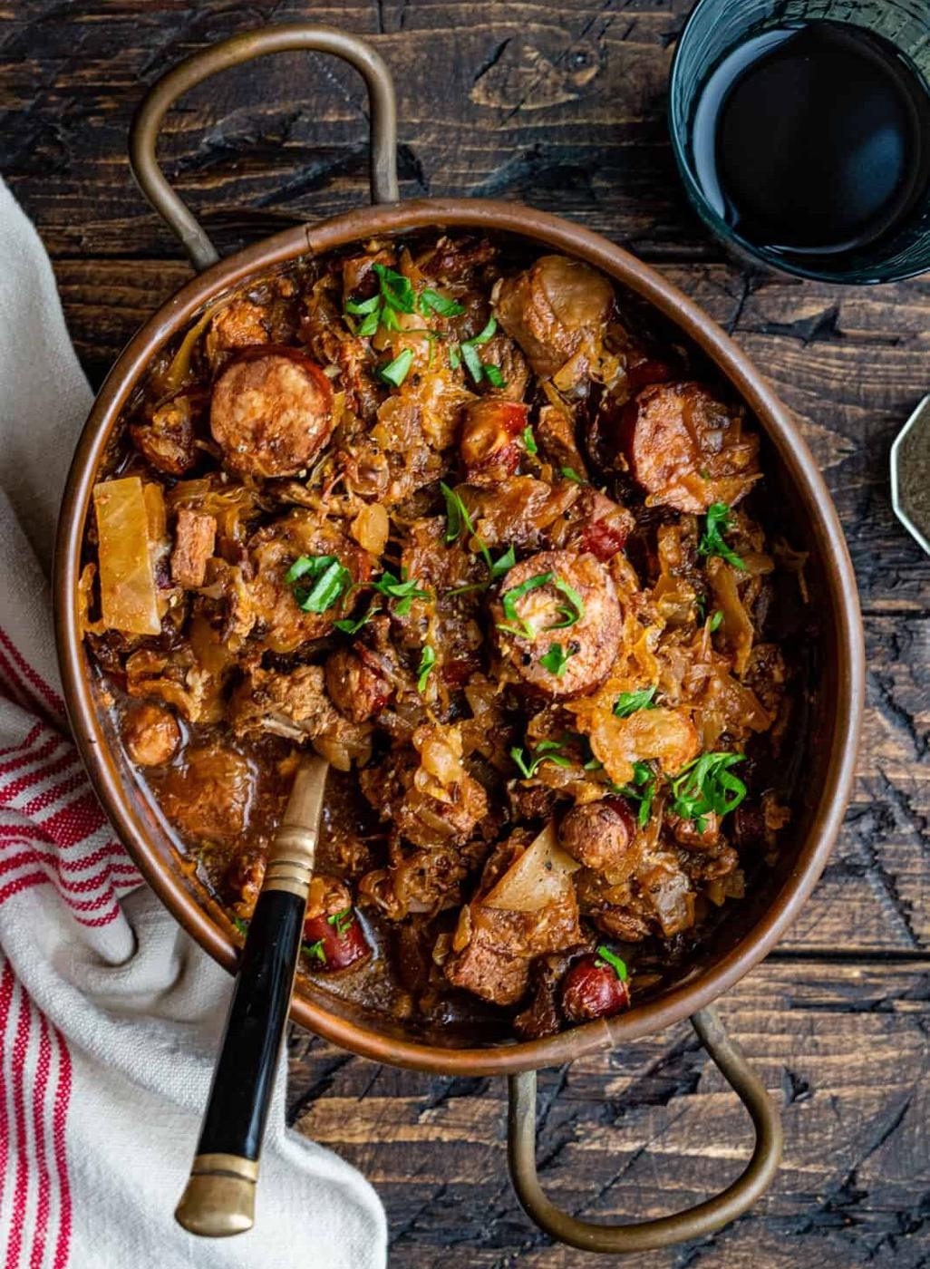 A rustic pot of Bigos Stew with sausages, meat, cabbage, and herbs sits on a wooden table next to a glass of dark beverage and a white towel with red stripes. A spoon rests in the pot.