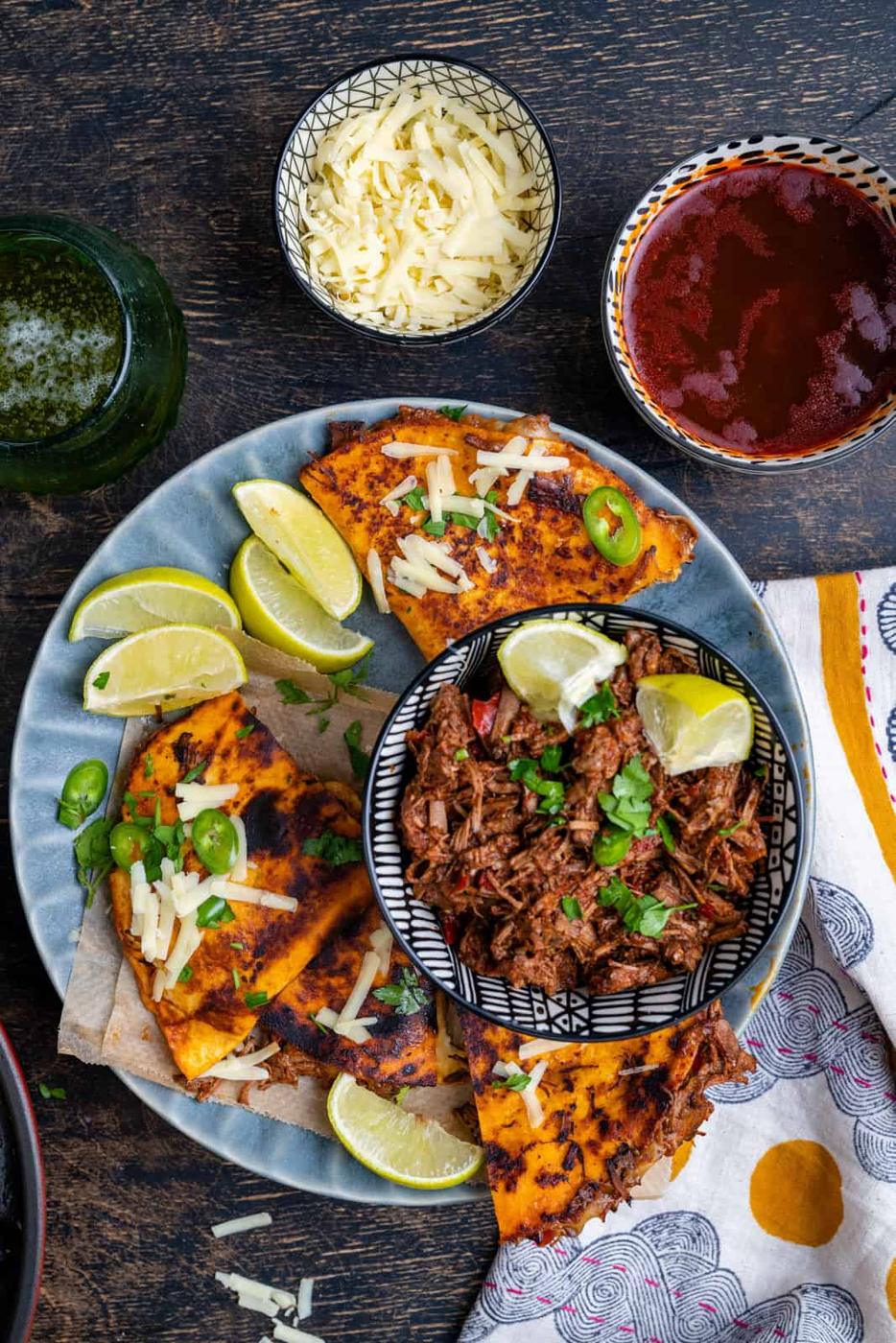 Birria tacos and birria stew on a plate with lime wedges and cheese on the side