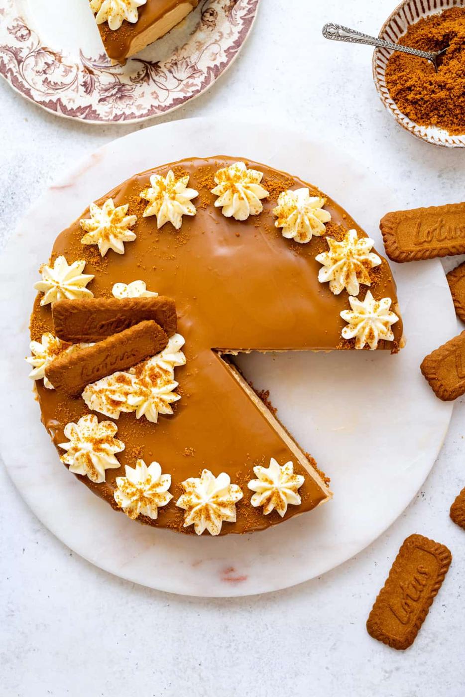 A round caramel-colored Biscoff no bake cheesecake topped with whipped cream, crumbled cookies, and two whole Lotus Biscoff cookies, with a slice removed. Nearby are extra cookies and a bowl of crumbs.