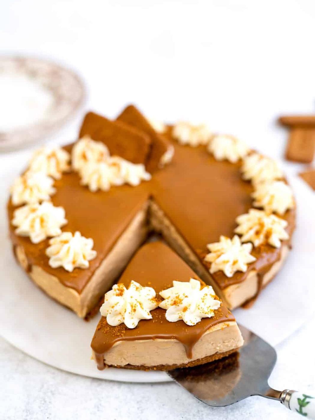 A caramel-colored Biscoff no bake cheesecake topped with whipped cream and cookie pieces, with one slice being lifted out on a cake server. The cheesecake sits on a white surface.