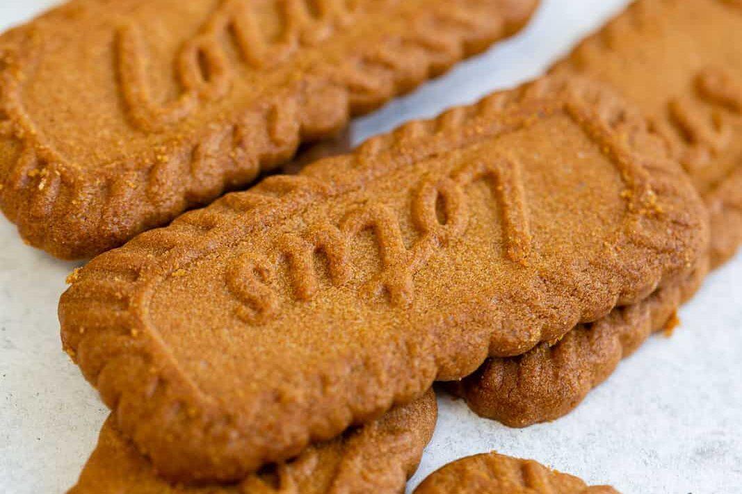 Several rectangular caramelized Lotus Biscoff cookies with scalloped edges and the word Lotus embossed on top, stacked on a light surface.