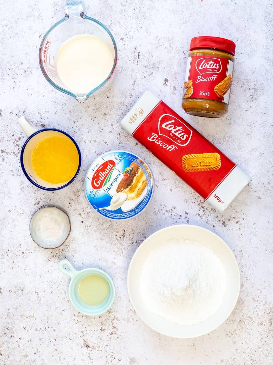 Assorted ingredients on a white surface: a jar of Lotus Biscoff spread, a package of Lotus Biscoff biscuits, a bowl of powdered sugar, a cup of melted butter, heavy cream, mascarpone cheese, lemon juice, and a small bowl of sugar.
