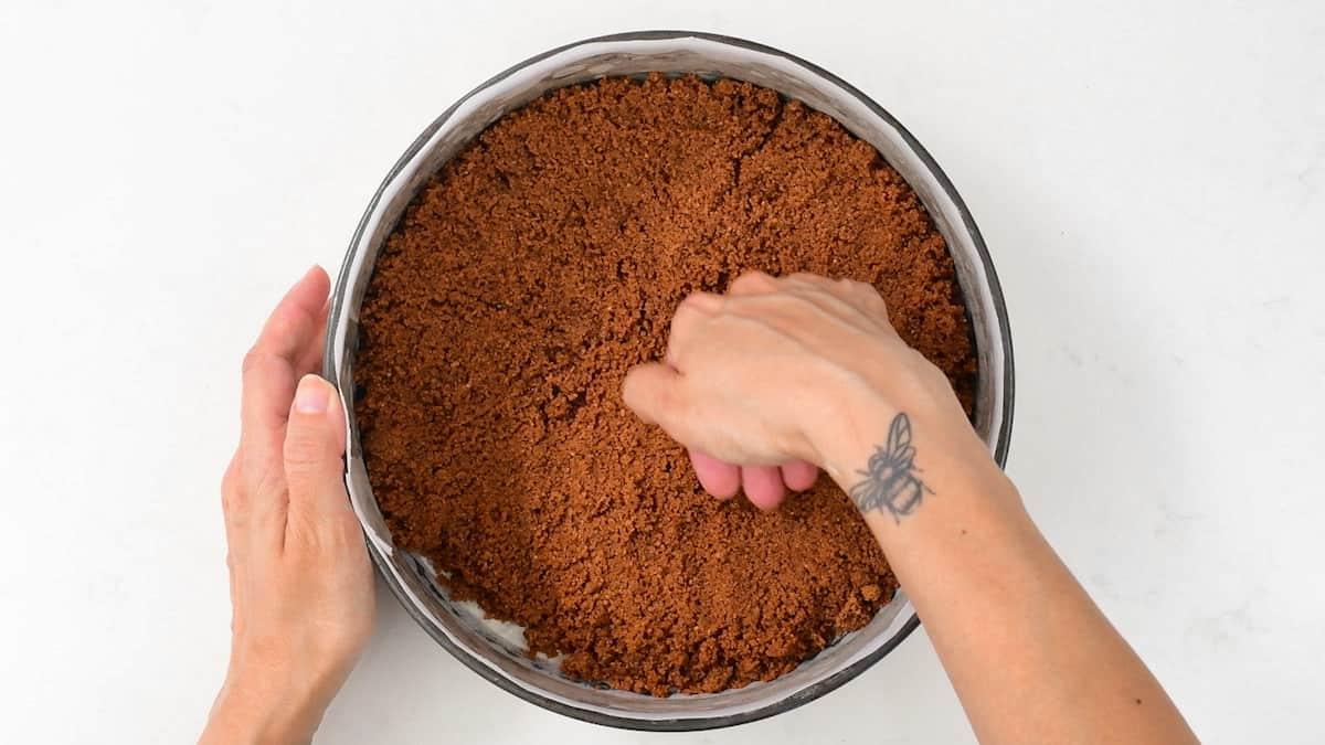 A person presses a crumbly Biscoff no bake cheesecake crust into a round baking pan. One hand holds the pan while the other presses down; a bee tattoo is visible on their wrist. The background features a white surface.