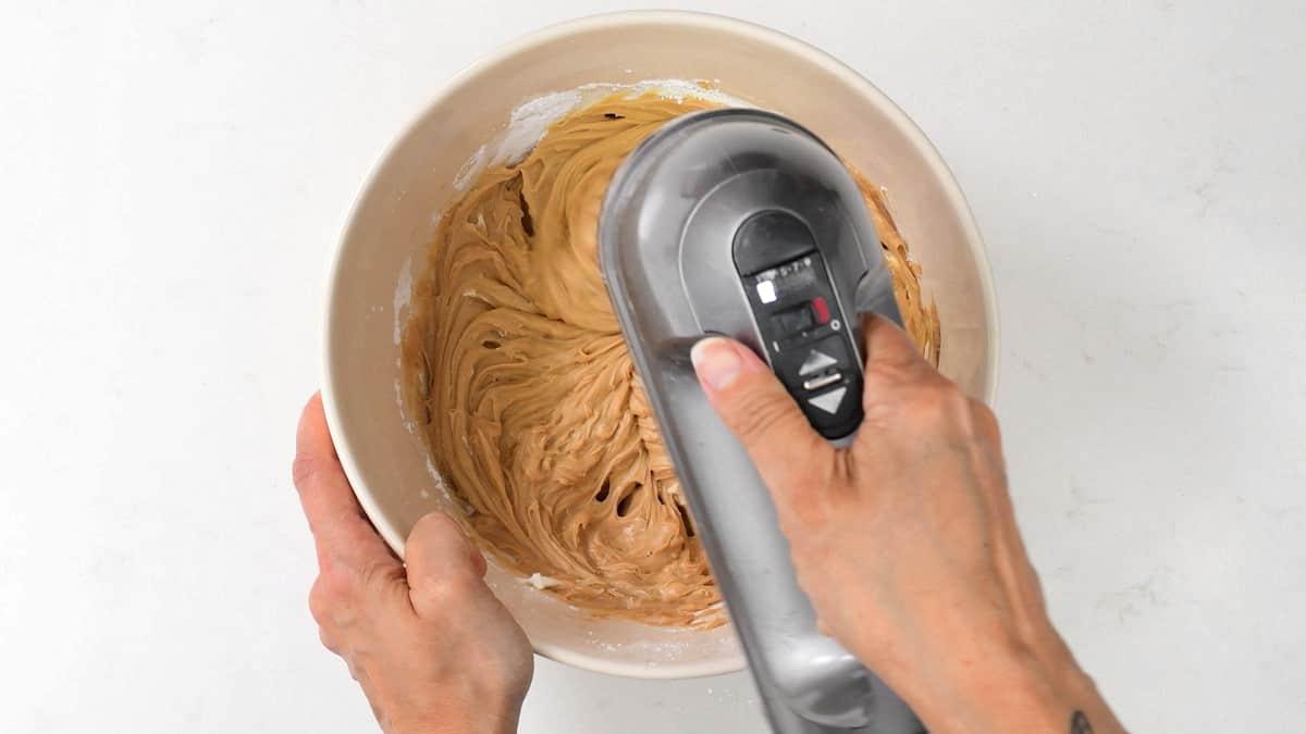 A person uses a hand mixer to blend light brown Biscoff no bake cheesecake batter in a white bowl on a white surface.
