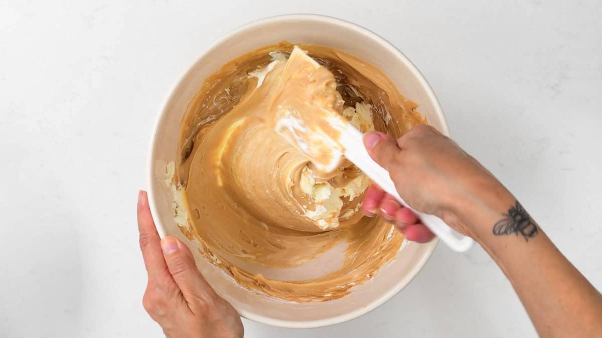 A person uses a white spatula to mix light brown and white ingredients for a Biscoff no bake cheesecake in a beige bowl. The person has a tattoo on their left wrist, and the bowl sits on a white surface.