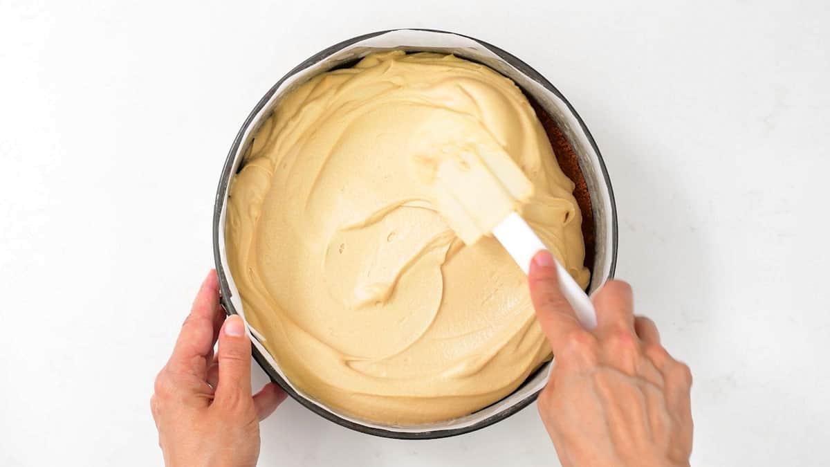 A person spreads creamy Biscoff no bake cheesecake batter evenly in a round baking pan using a white spatula, viewed from above against a white background.
