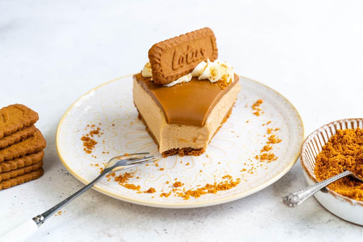 A slice of Biscoff no bake cheesecake topped with a Lotus Biscoff cookie and whipped cream sits on a decorative plate, with a fork, extra cookies, and a bowl of cookie crumbs beside it.