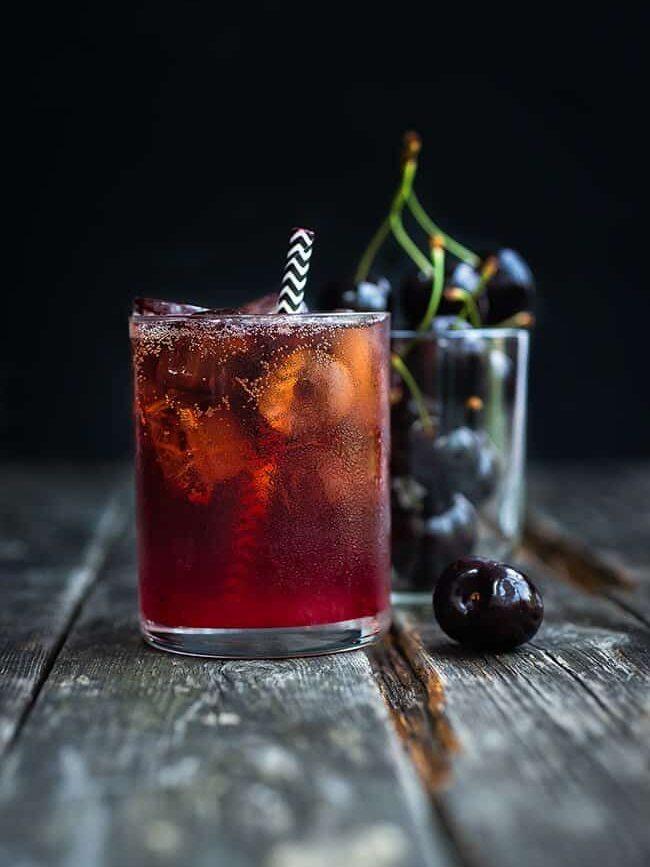 A glass of iced Black Cherry Bourbon Cola Smash cocktail with a striped straw sits on a rustic wooden surface, while another glass filled with fresh cherries is in the background.