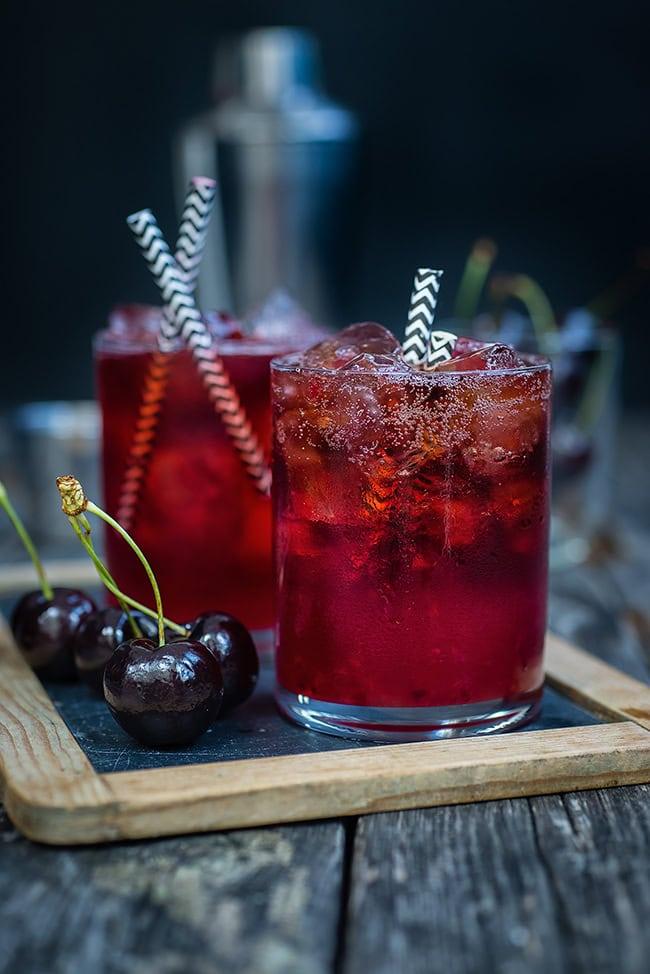 Two glasses of iced black cherry cola smash cocktails with black and white striped straws sit on a wooden tray beside fresh cherries, with a cocktail shaker blurred in the background.
