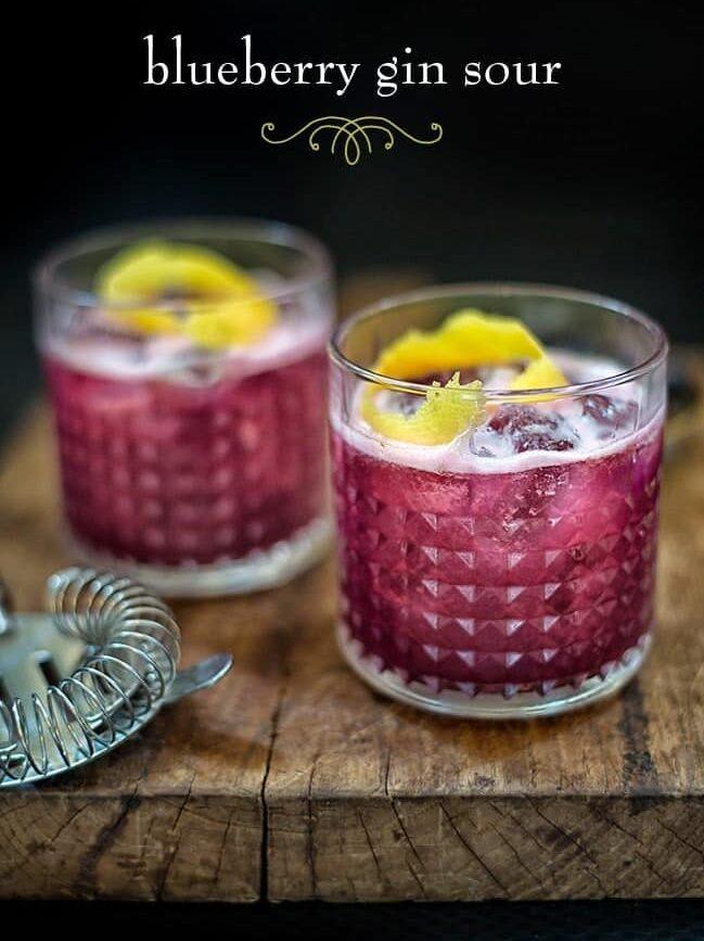 Two glasses of blueberry gin sour with ice and lemon peels sit on a rustic wooden board, accompanied by a metal cocktail strainer. The text "Blueberry gin sour" is displayed at the top, highlighting this refreshing cocktail.