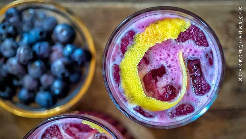 A close-up of a blueberry gin sour cocktail—pink, frothy, and served with ice cubes and a twist of lemon peel—alongside a bowl of fresh blueberries on a wooden surface.