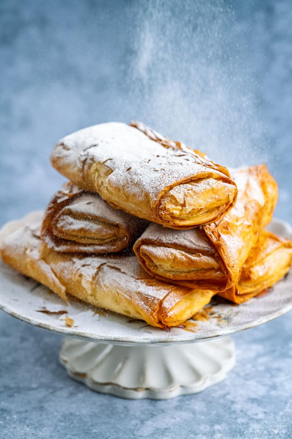 Bougatsa pastries being dusted with icing sugar and cinnamon on a cake stand