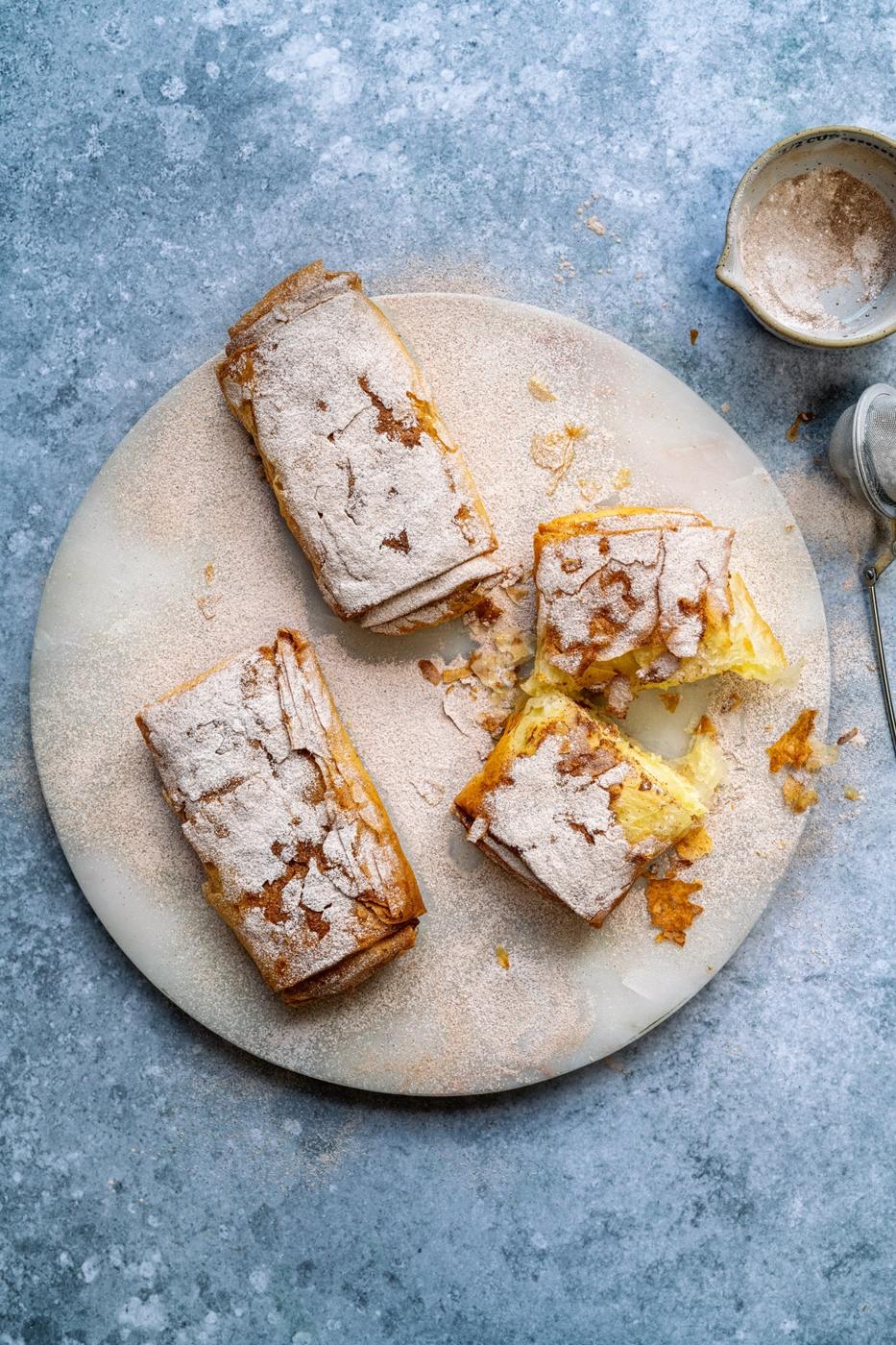 Greek Bougatsa, three individual pastries dusted with icing sugar and cinnamon on a marble board, one of the pastries cut in half