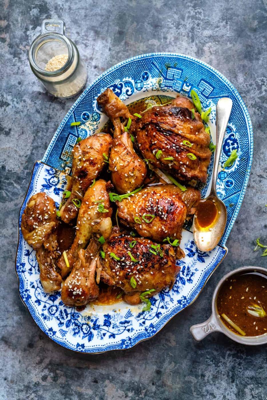 A blue and white platter holds glazed braised chicken thighs garnished with green onions and sesame seeds, with a spoon, a small jug of sauce, and a jar of sesame seeds nearby—all set on a textured gray surface.