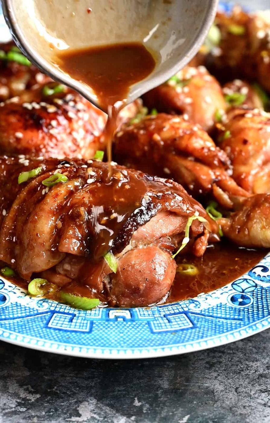 A close-up of Braised Chicken Thighs on a blue-patterned plate, with brown sauce being poured over them. The chicken is garnished with sesame seeds and chopped green onions, hinting at the rich flavors from the Dutch Oven.