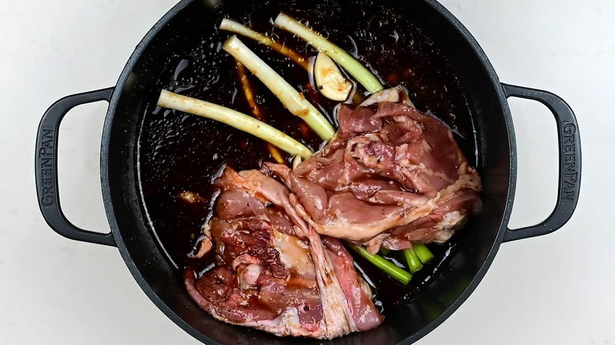 Braised Chicken Thighs, green onions, and garlic rest in a dark marinade inside a black Dutch Oven on a white surface, ready for cooking.