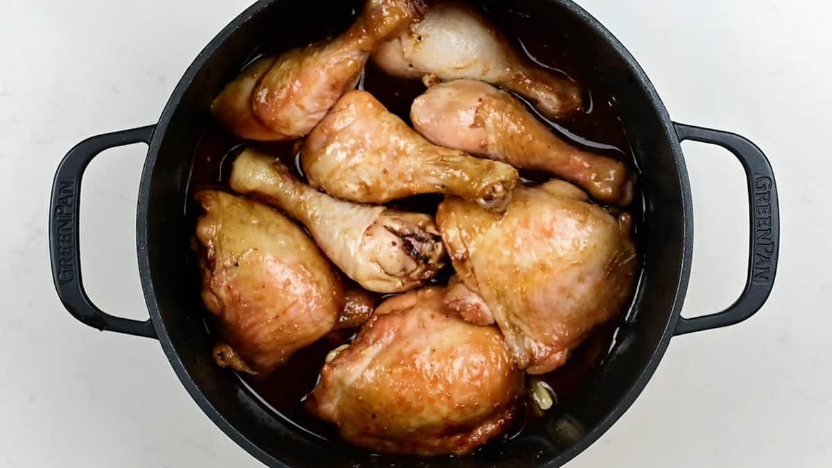 A black Dutch Oven filled with braised chicken thighs and drumsticks in a rich brown sauce, seen from above on a white surface.