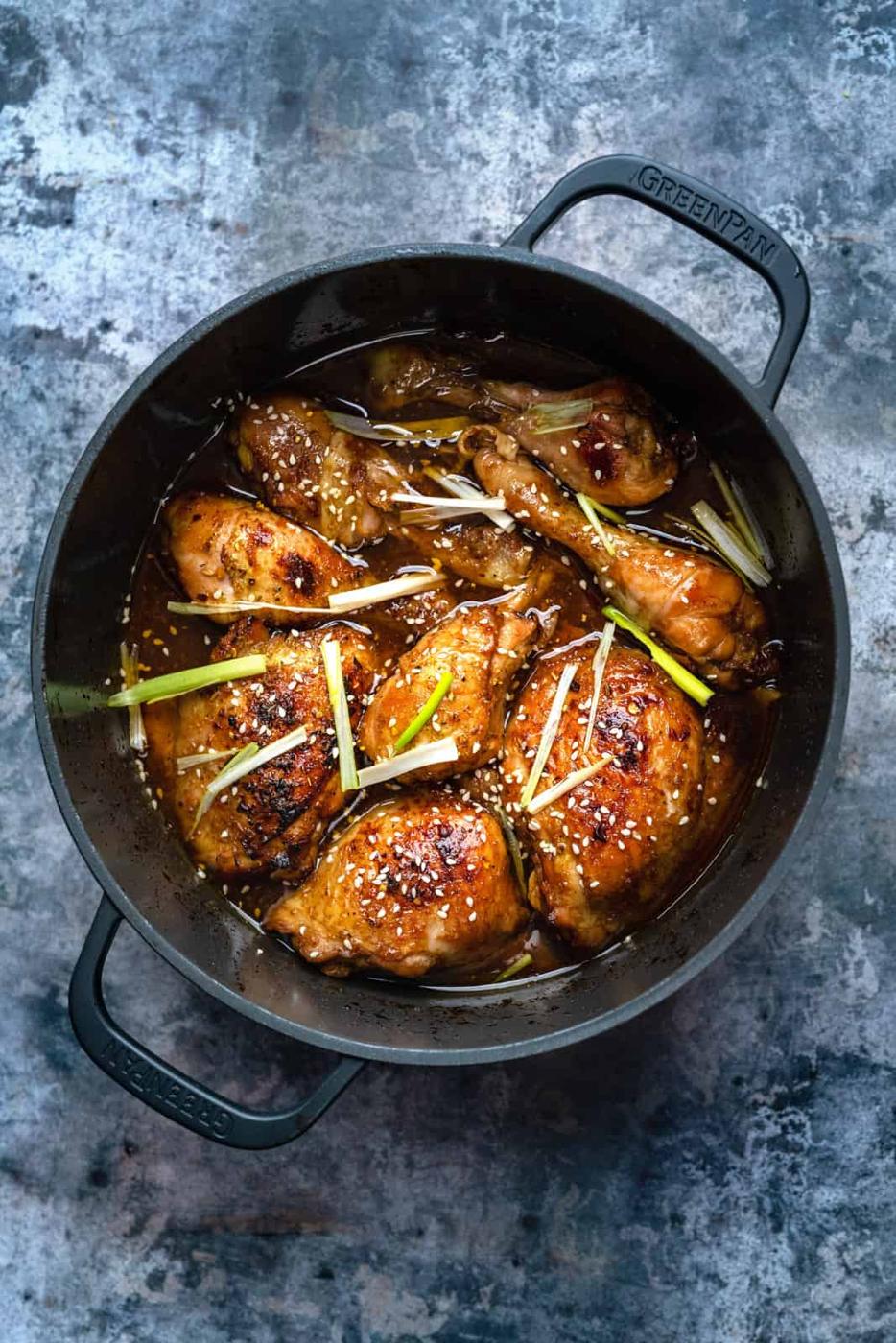 A black Dutch Oven filled with glazed, browned braised chicken thighs, garnished with sliced green onions and white sesame seeds, sits on a textured gray surface.