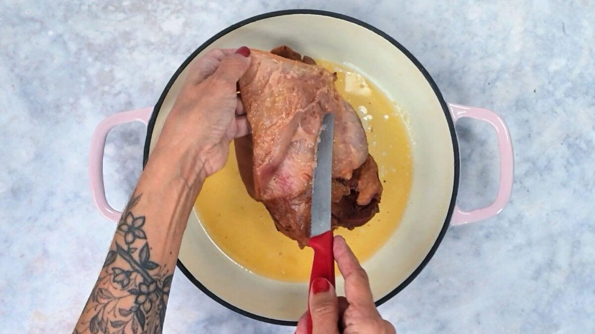 Peeling the rind off a braised ham hock with a small knife, held over the pot.
