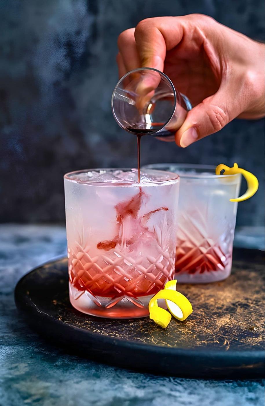 A hand pours crème de mûre over a Bramble cocktail in a glass of crushed ice so it bleeds through the drink, with a lemon twist garnish.